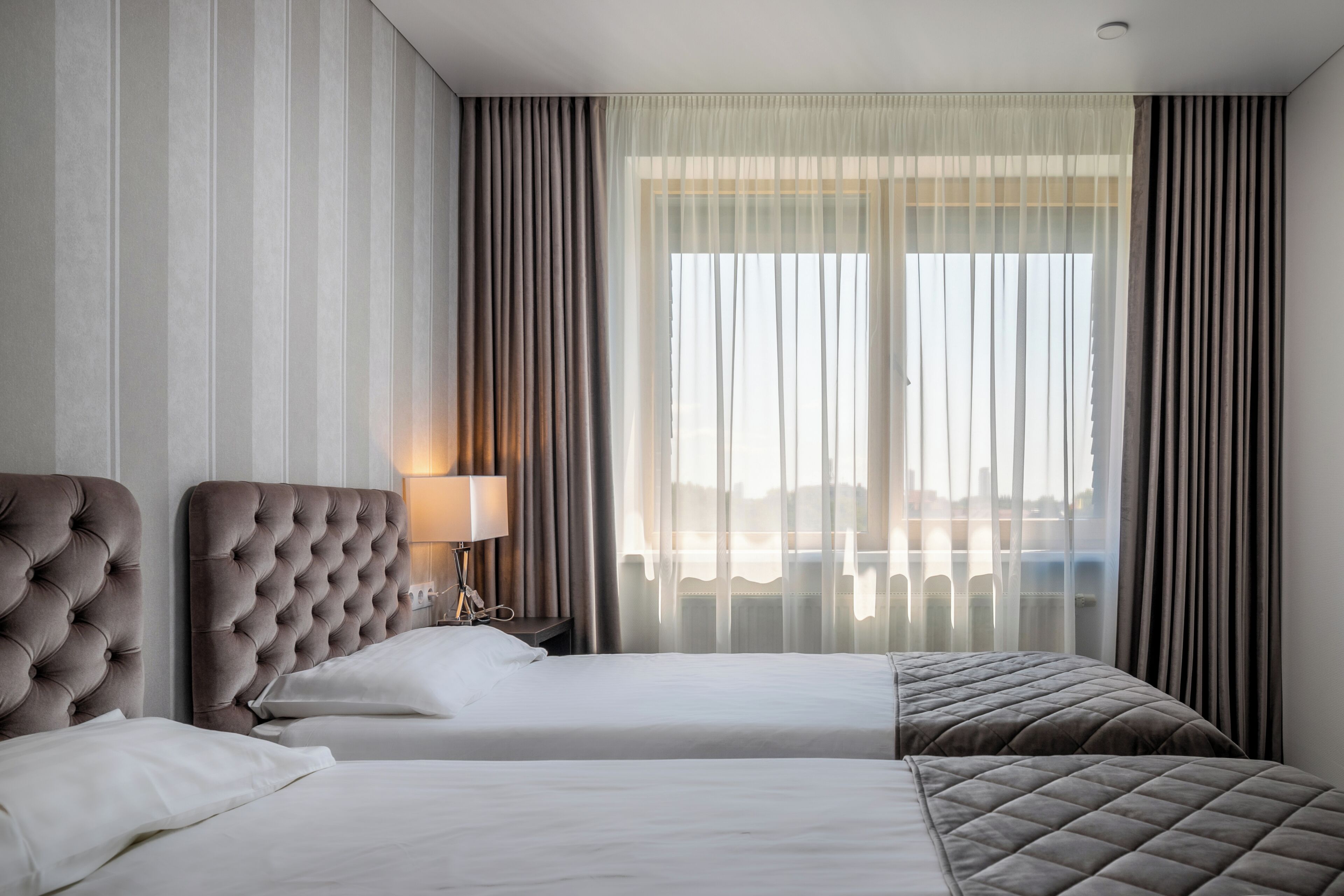 A hotel room with two beds, a large window with curtains, a lamp, and a view of the cityscape.