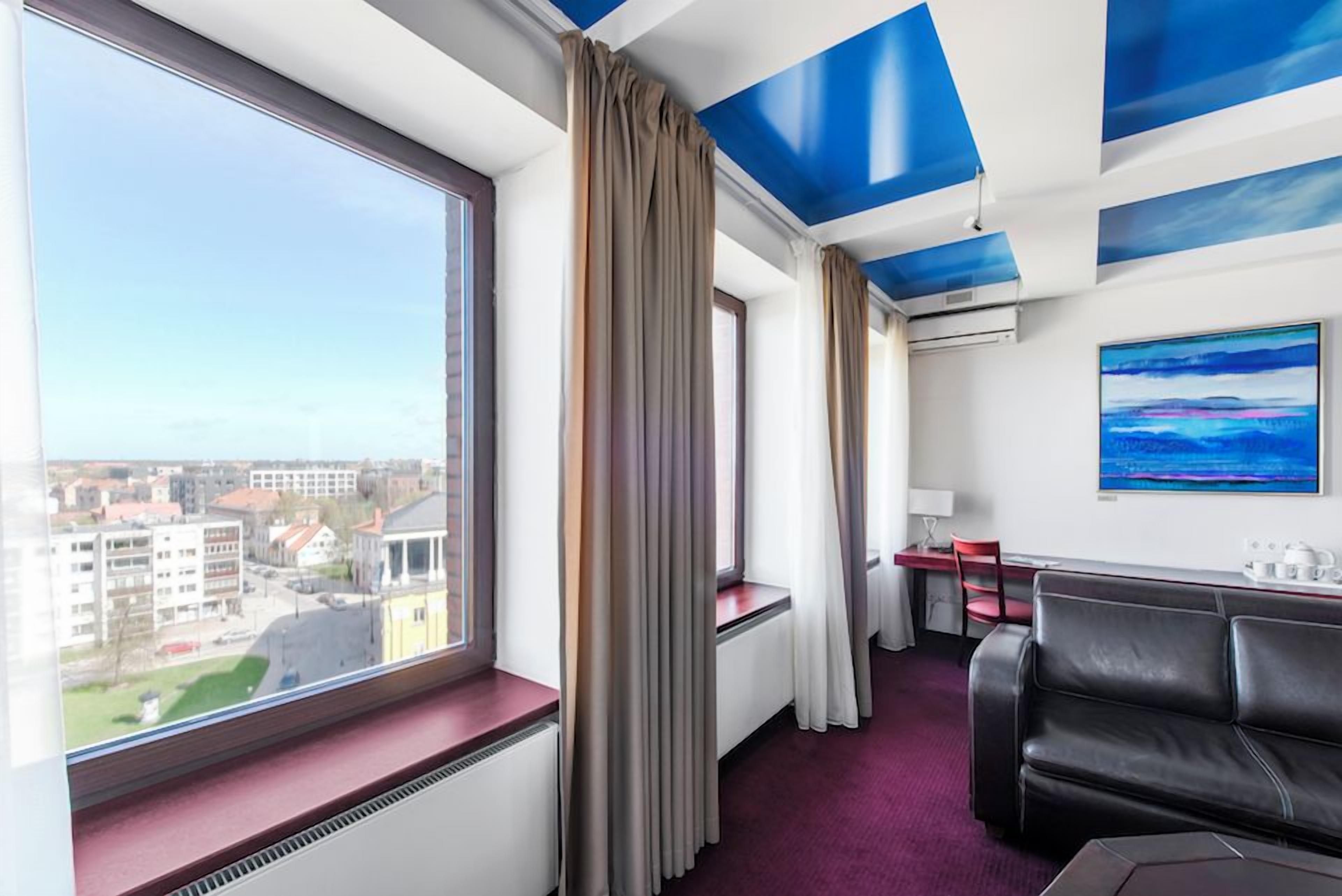 Ein Hotelzimmer mit großem Fenster, einer schwarzen Ledersofa, einem Schreibtisch mit Stuhl und einem Bild an der Wand.
