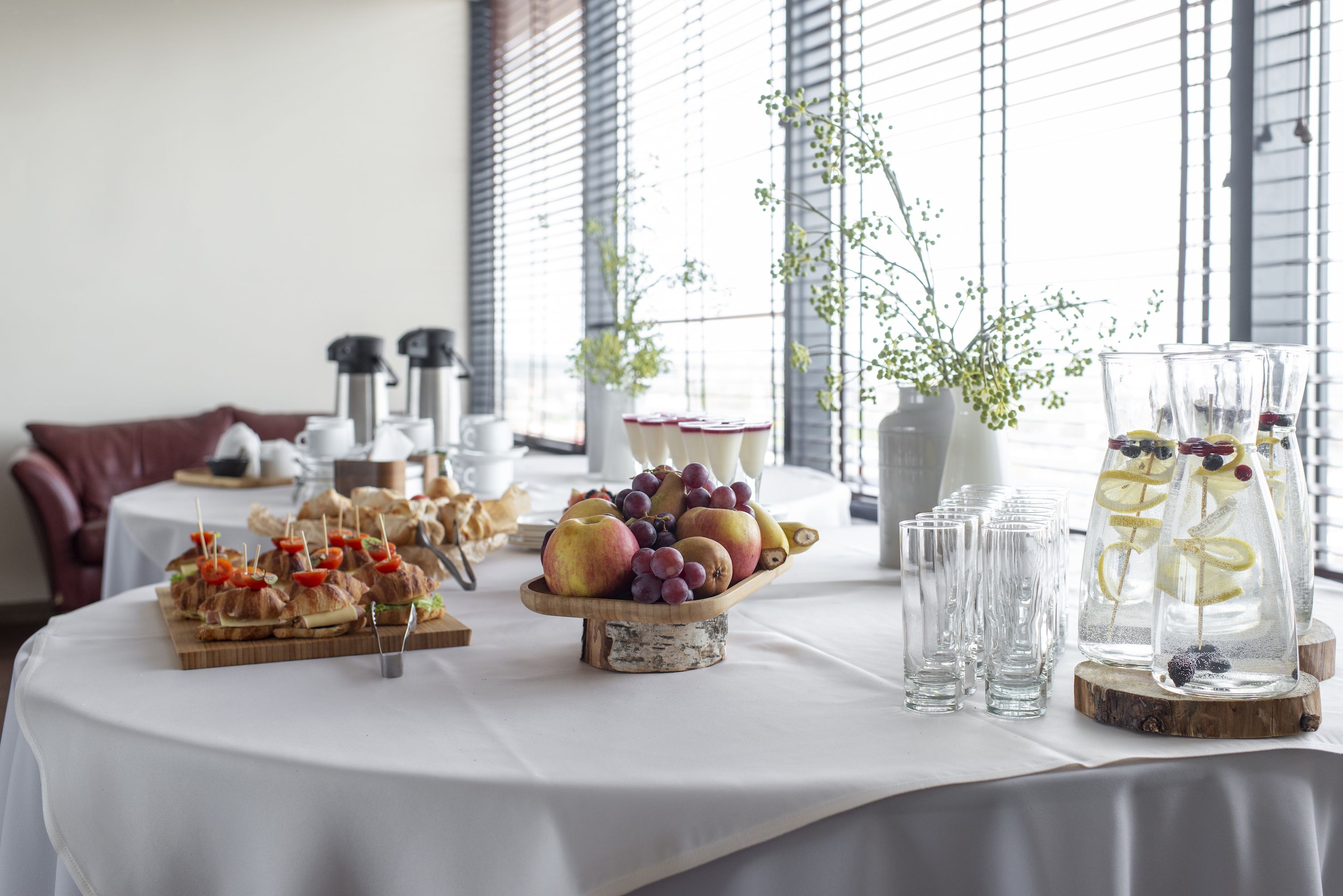 Ein Tisch mit Speisen und Getränken, eine Vase mit Blumen und ein Fenster mit Jalousien.