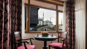 A room with a view of a mosque and minaret through a window, a round table with chairs, and a basket of fruit on the table. - Seven Hills Hotel - Special Class (Istanbul)