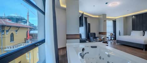 A modern hotel room with a large bathtub, a bed, a seating area with chairs, and a view of the cityscape.