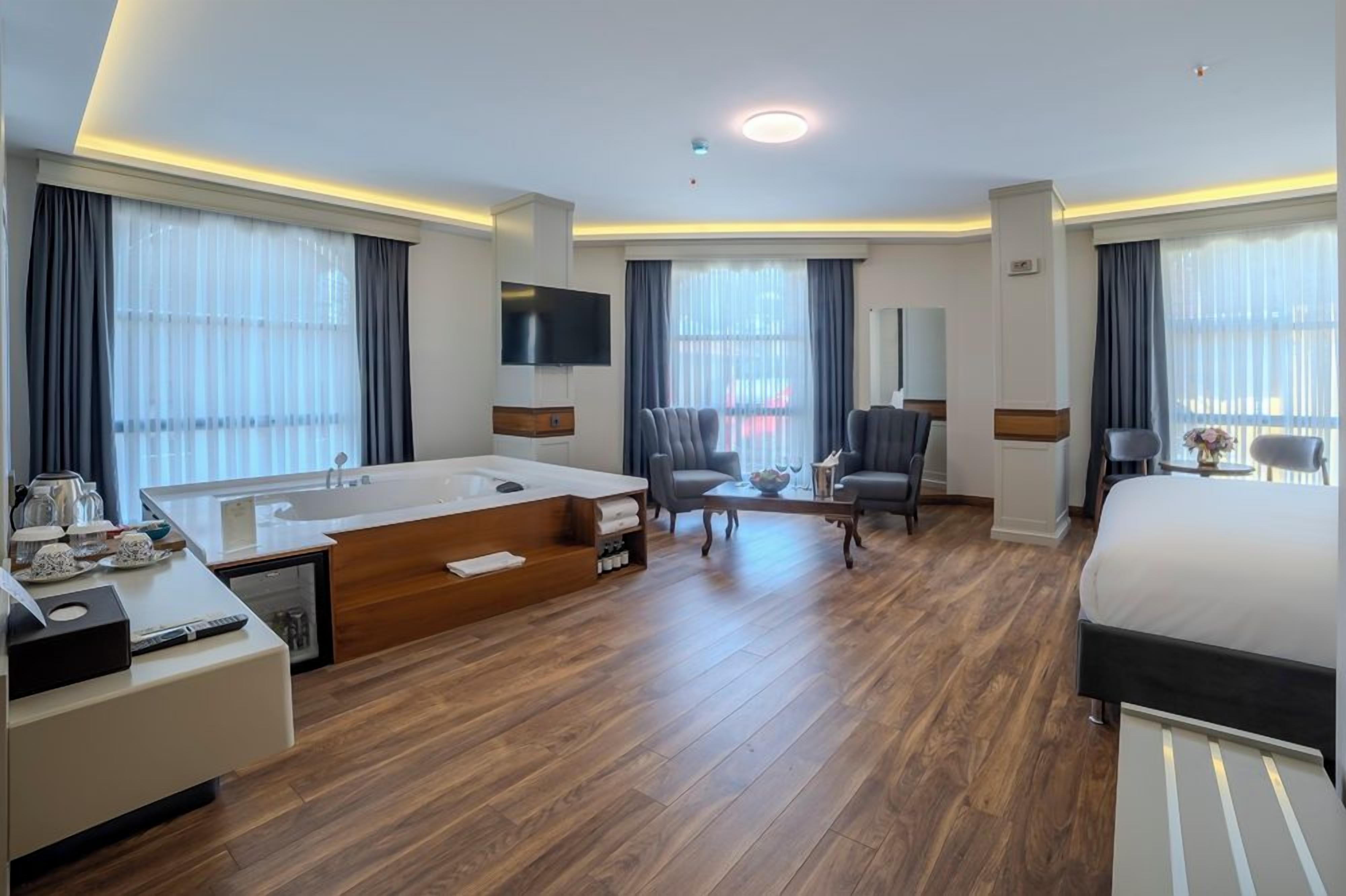 A modern hotel room with a large bed, a separate sitting area with a sofa and armchairs, a small table, a television, and a large window with curtains.
