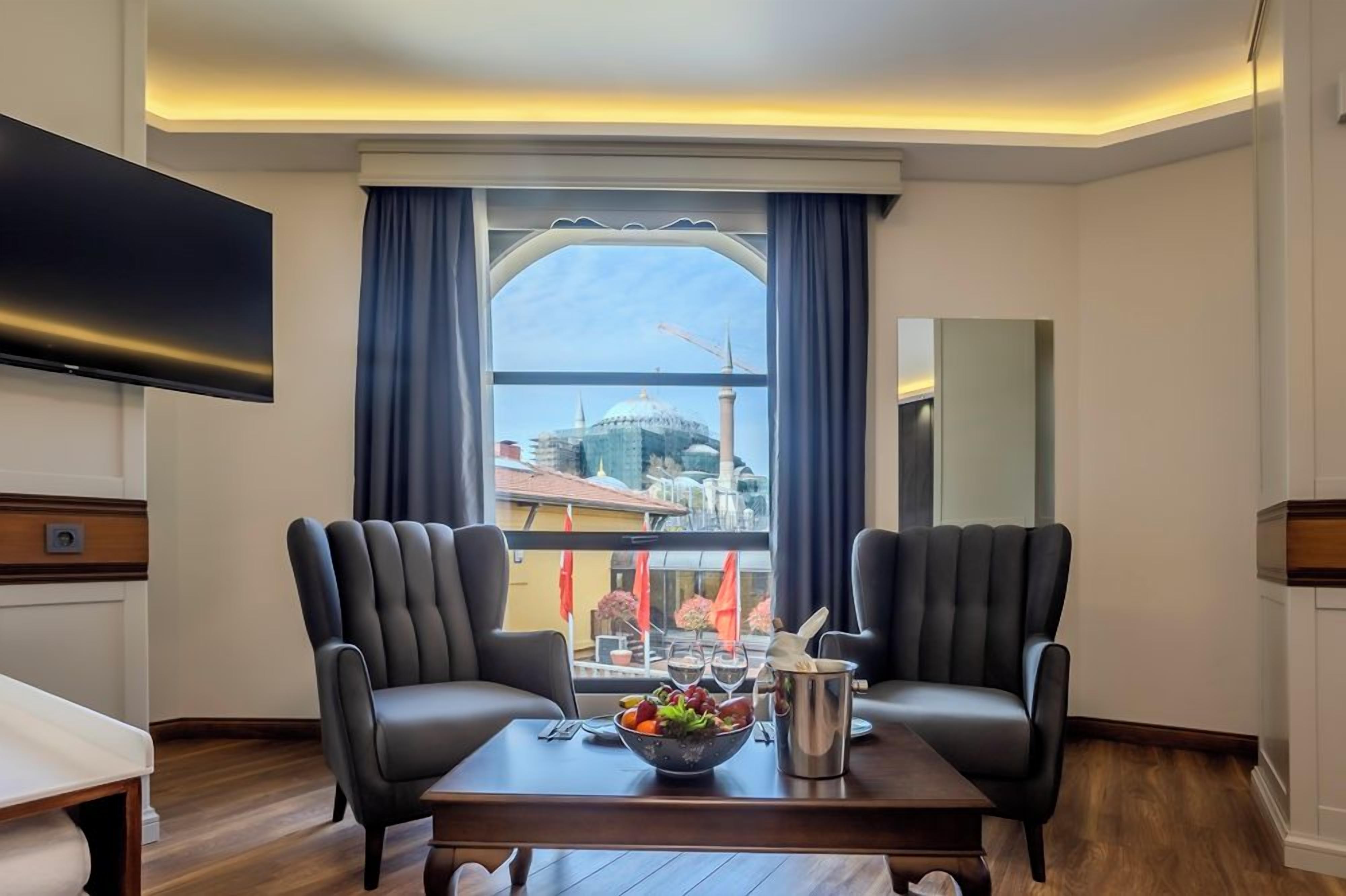 Ein Hotelzimmer mit Fernseher, zwei Ledersesseln, einem Esstisch mit Obstschale und Blick auf eine Stadtlandschaft durch ein Fenster.