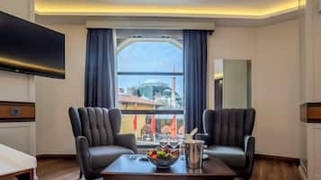 Ein Hotelzimmer mit Fernseher, zwei Ledersesseln, einem Esstisch mit Obstschale und Blick auf eine Stadtlandschaft durch ein Fenster.