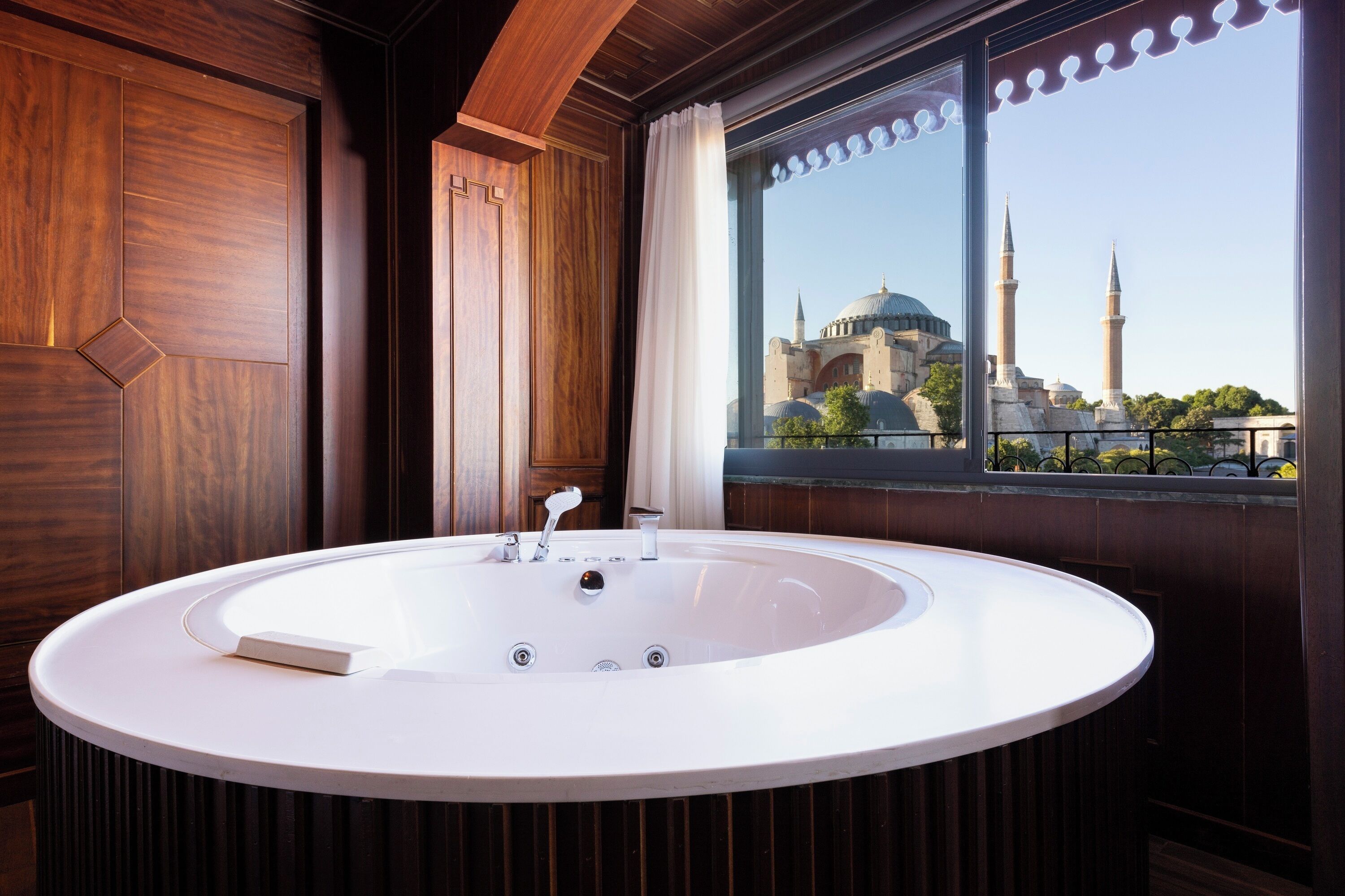 A modern bathroom with a large oval bathtub, wooden paneling, and a view of a mosque through a large window.