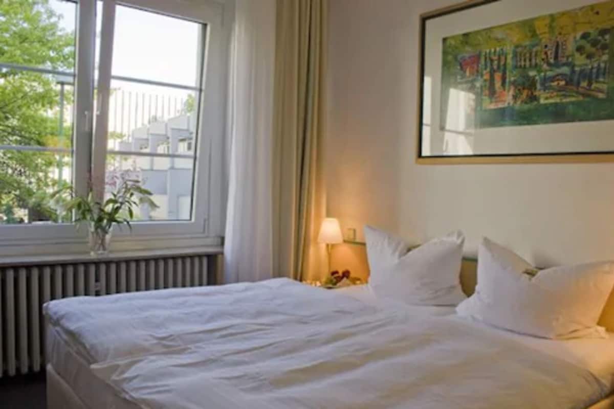 A hotel room with a bed, a window with curtains, a radiator, a nightstand with a lamp, and a framed painting on the wall.