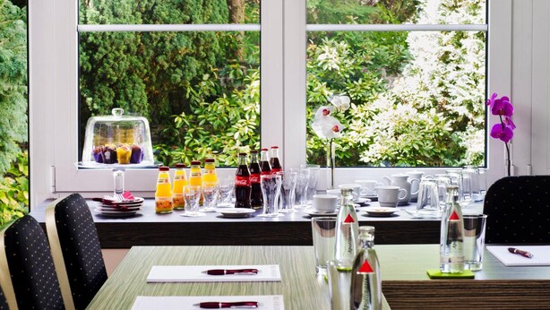 A breakfast setup with a variety of drinks, a fruit display, and a view of lush greenery through large windows.