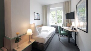 A hotel room with a bed, bedside table, lamp, desk, chair, TV, and a window with curtains.