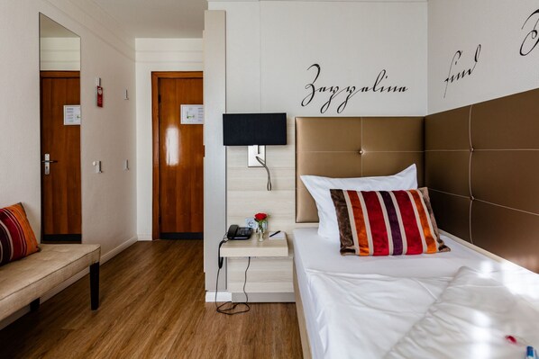 Habitación de hotel con suelo de madera, cama con almohadas blancas y a rayas, mesita de noche con lámpara, pequeño florero con una rosa y pared con el texto 'Zanzibar fin'.