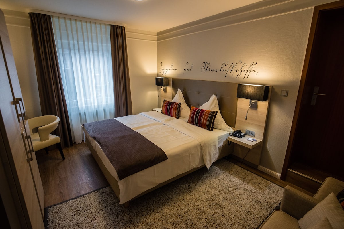 A hotel room with a large bed, two bedside tables, a desk, a chair, and a large window with curtains.