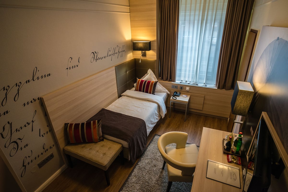 A hotel room with a bed, a desk, a chair, a nightstand, a lamp, and a window with curtains.
