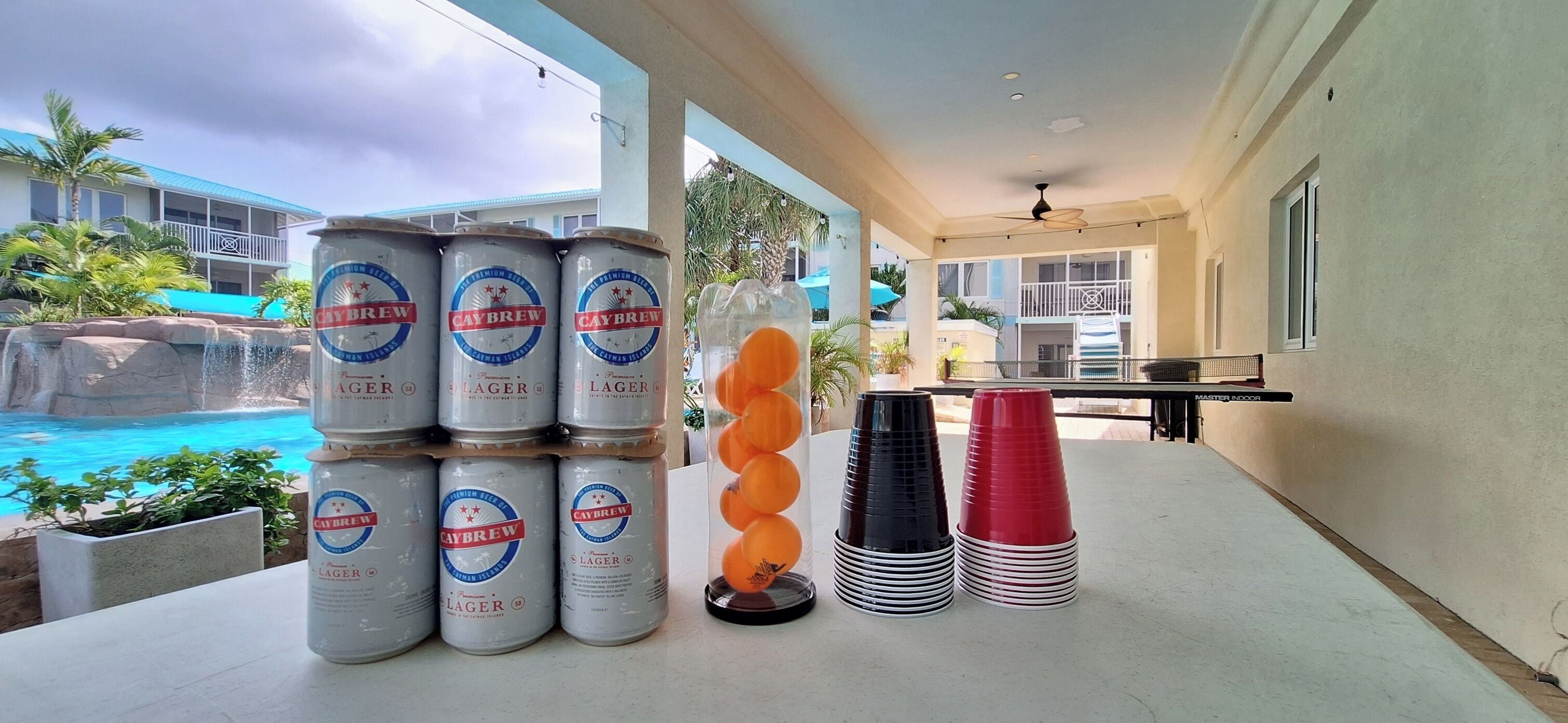 Zona de piscina con latas de cerveza apiladas, vasos de plástico y vista a un edificio con balcón.