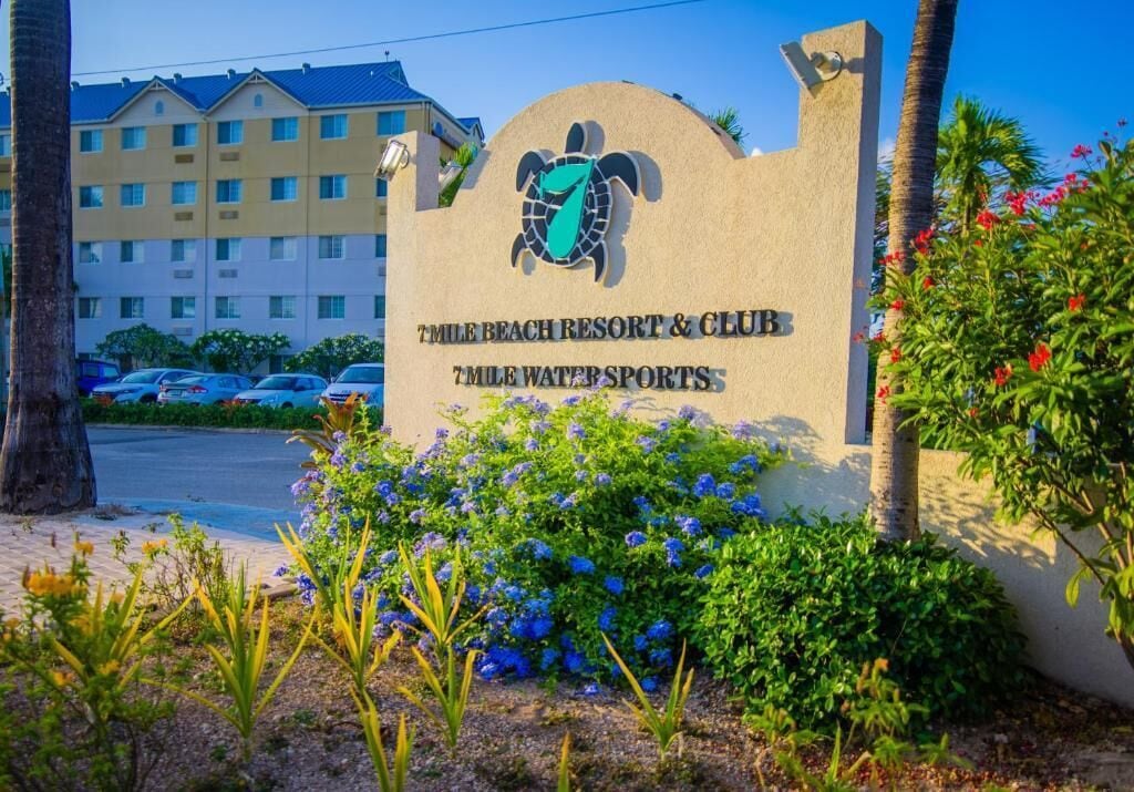 Señal de 7 Mile Beach Resort & Club, con logotipo de tortuga, rodeada de follaje y un edificio al fondo.