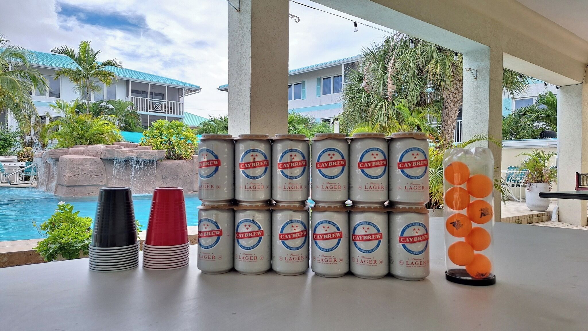 Un espace aménagé au bord de la piscine, avec des piles de canettes de bière, des gobelets en plastique et un vase décoratif contenant des sphères oranges.