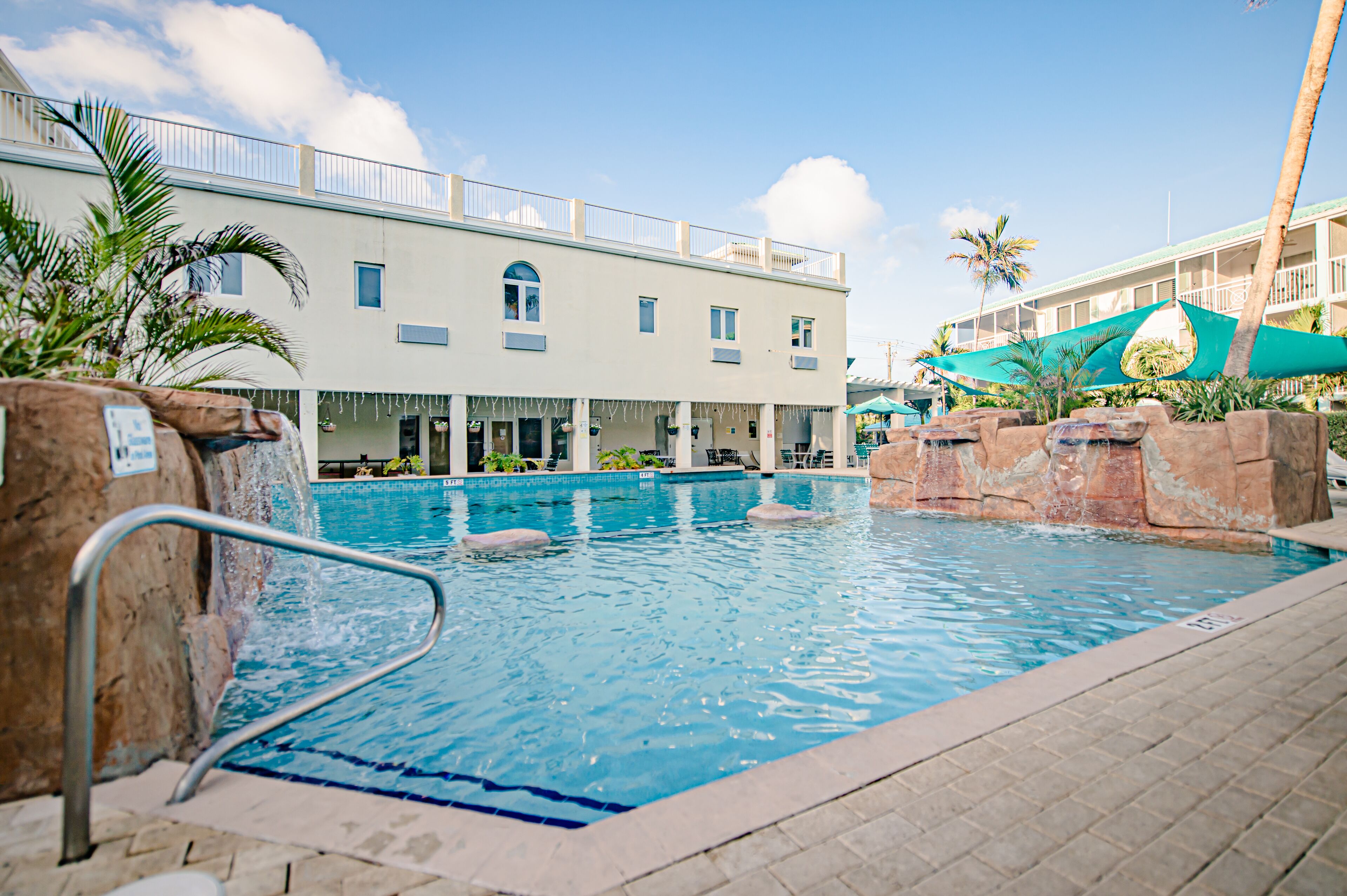 Una piscina con borde de piedra, escalera metálica y un edificio con balcón al fondo.