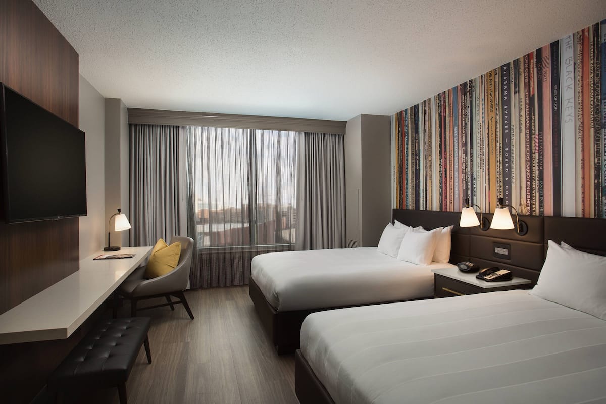 A hotel room with two beds, a desk, a chair, a TV, and a bookshelf with books.