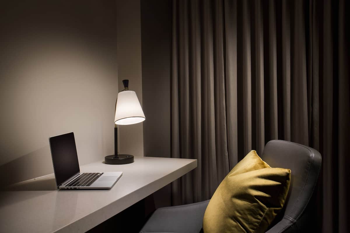 A laptop on a desk with a lamp and a chair with a pillow.