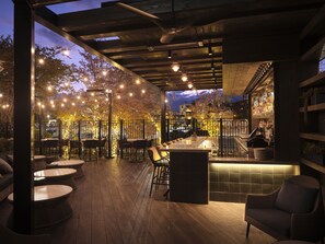 Un espacio al aire libre con mesas y sillas, una zona de bar con mostrador y vistas a la ciudad al atardecer.