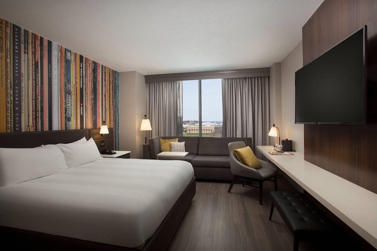 A hotel room with a large bed, a sofa, a desk, and a TV. There is a window with curtains and a view of buildings outside.