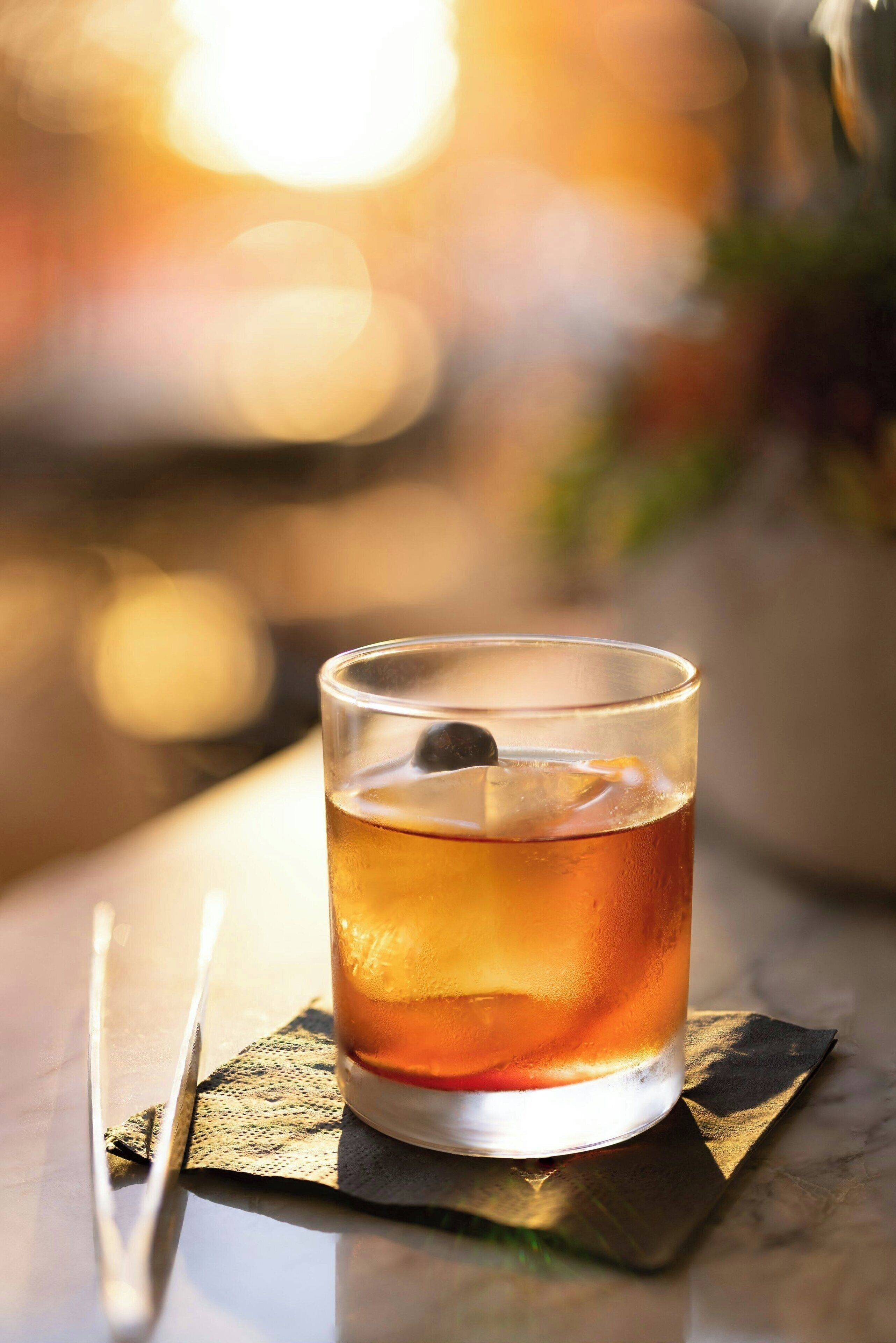 A glass of amber-colored beverage on a table with a napkin and a spoon.