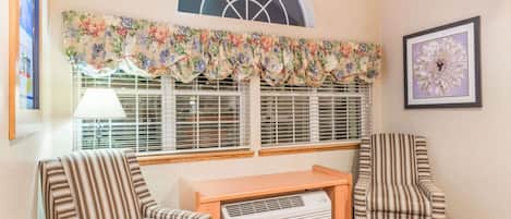 A room with two striped armchairs, a small wooden table, a window with a floral valance, and a framed artwork on the wall.