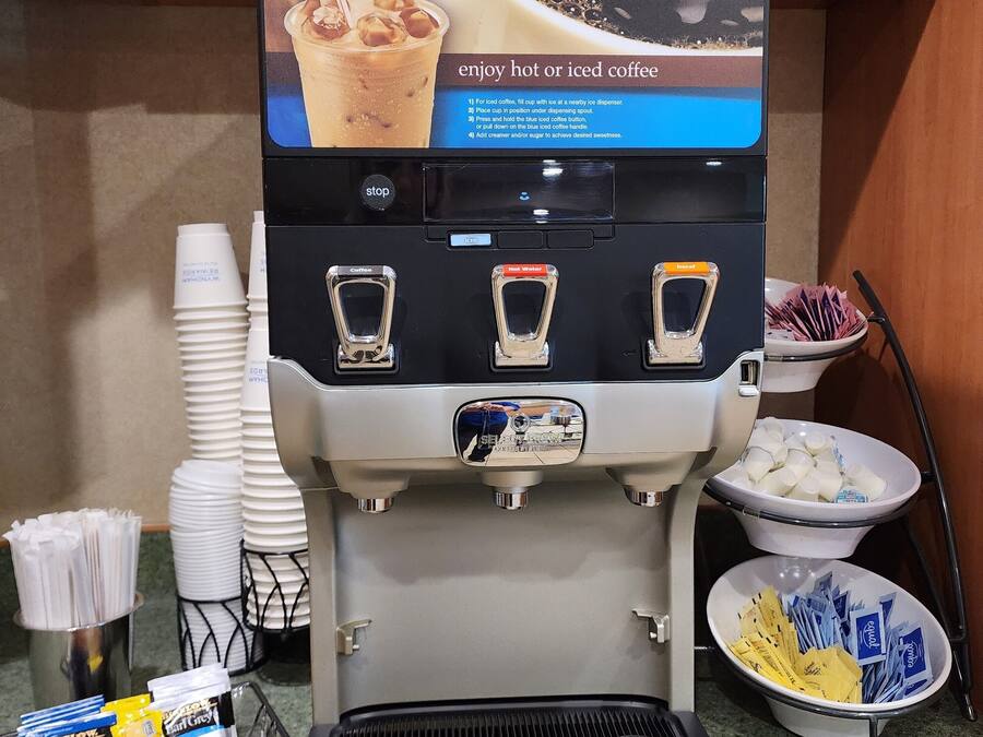 A coffee machine with a Folgers coffee cup and instructions, a coffee dispenser, and a tray with disposable cups and condiments.