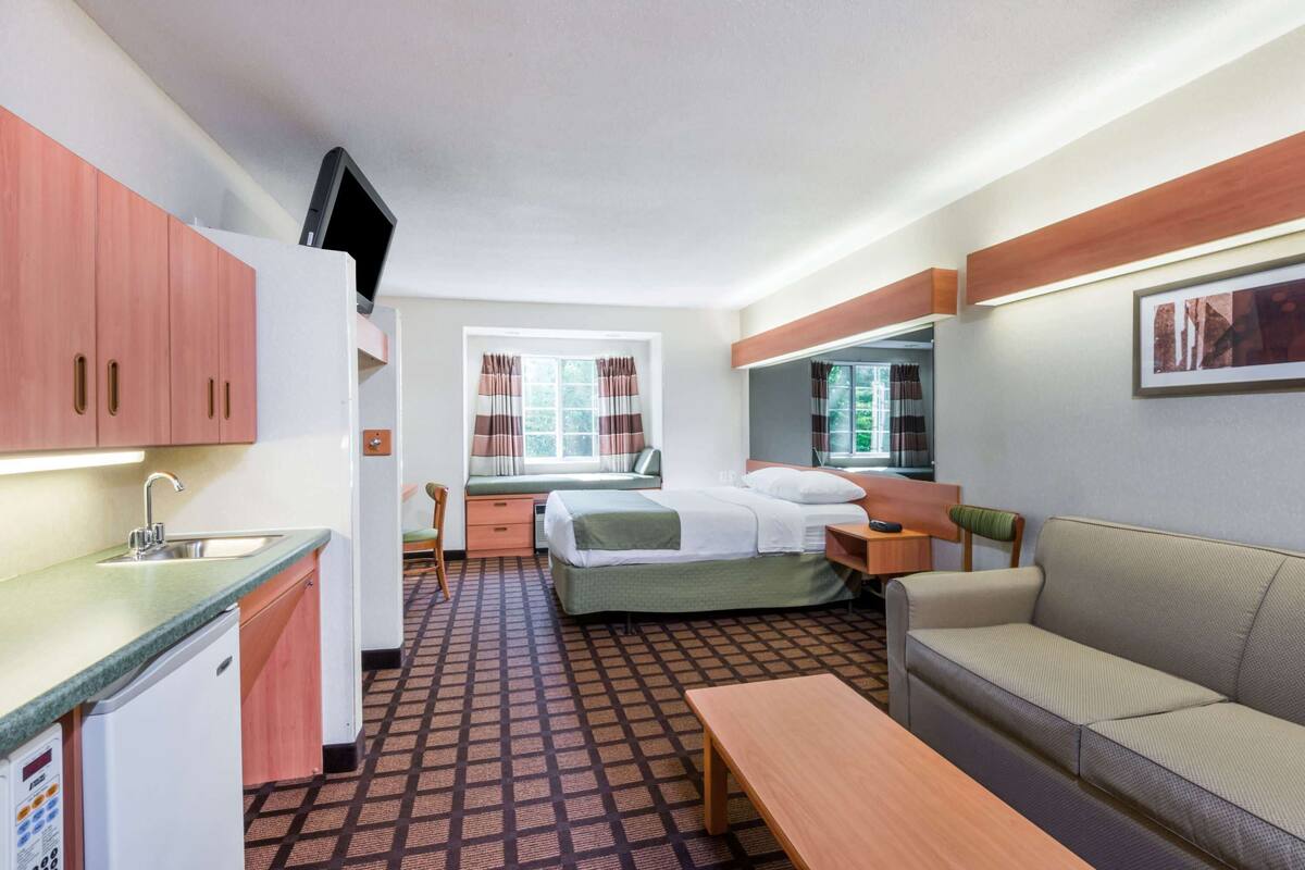 A compact hotel room with a kitchenette, a bed with a green bedspread, a sofa, a bedside table, and a window with curtains.