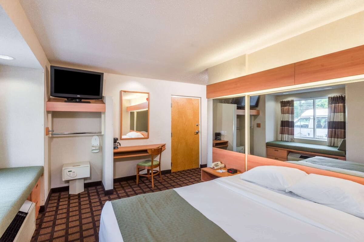 A hotel room with a bed, a desk with a chair, a television, a mirror, a window with curtains, and a small table with a phone and a lamp.
