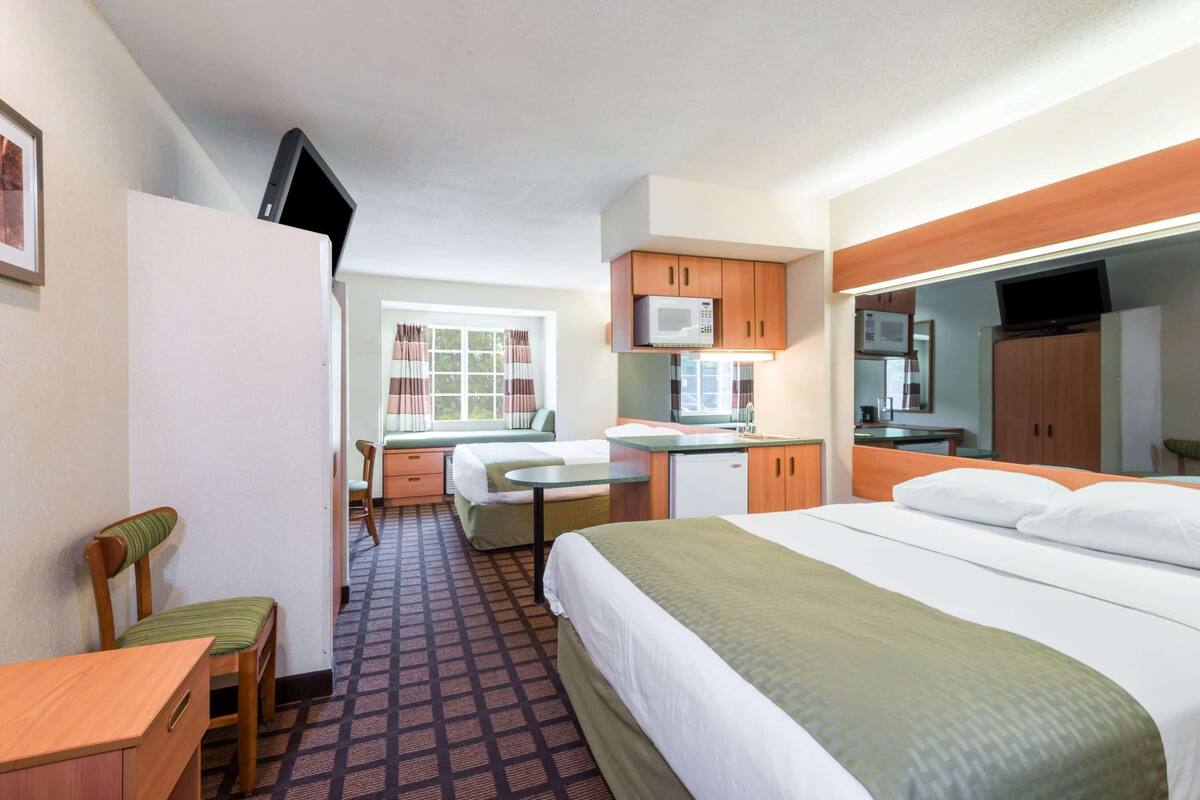 A hotel room with a bed, a desk, a chair, a kitchenette, and a television.