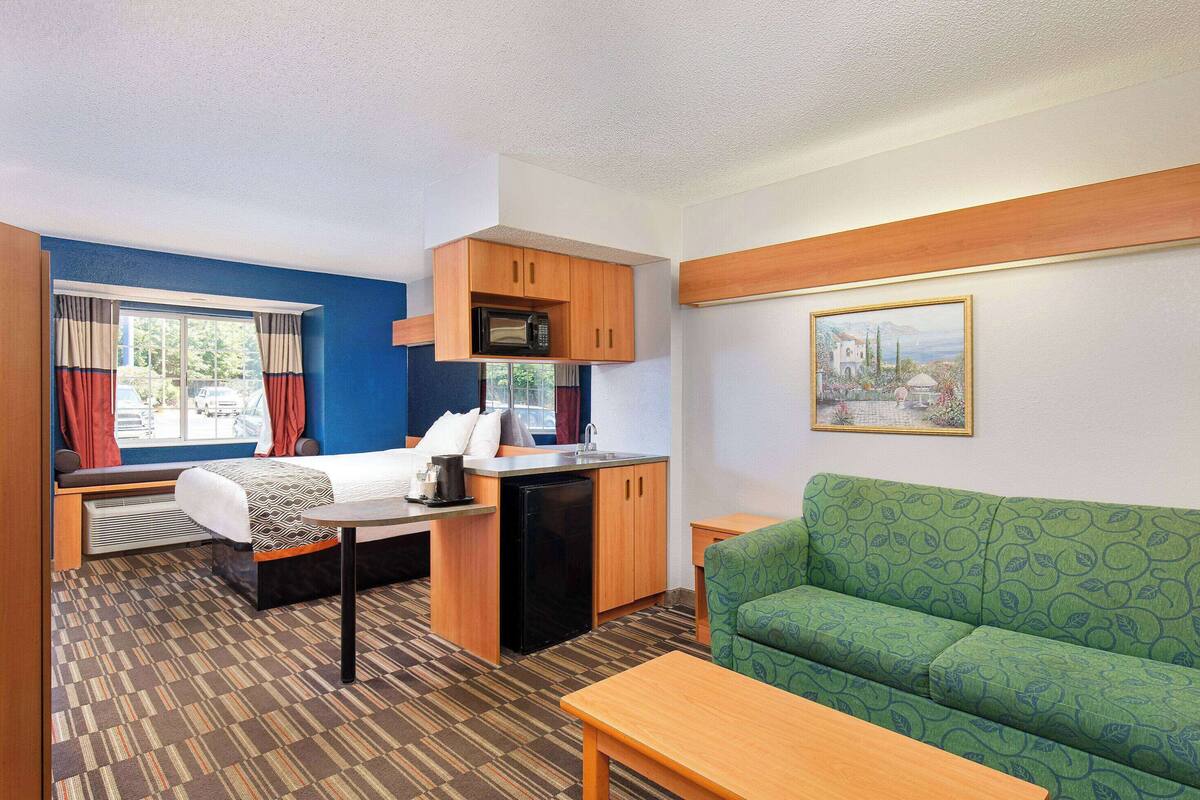 A compact room with a kitchenette, a green patterned sofa, a small table, and a window with curtains.