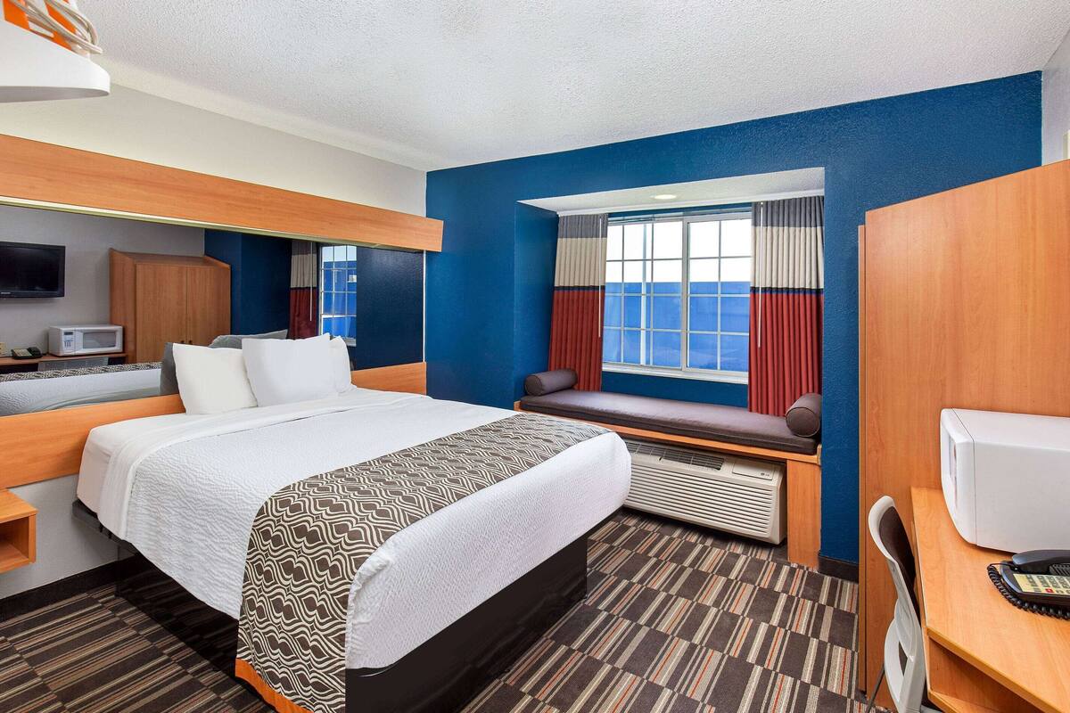 A hotel room with a large bed, a desk with a computer, a TV, and a window with curtains.