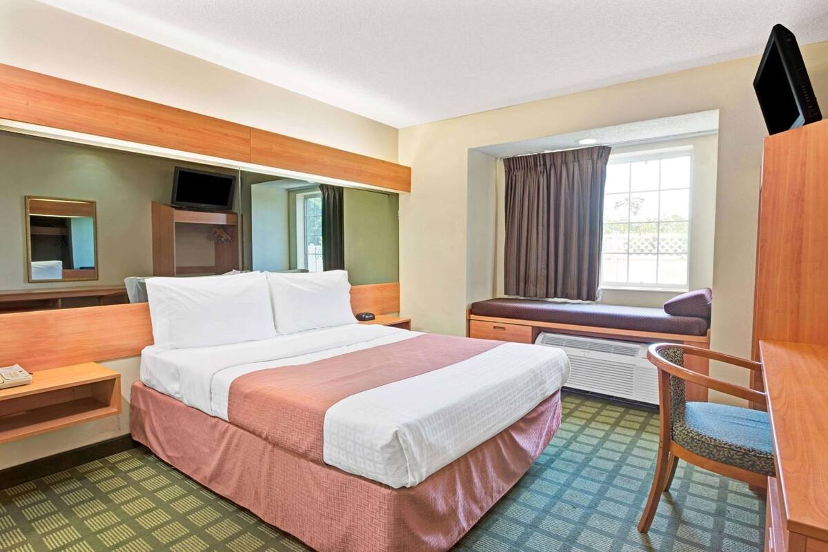 A hotel room with a bed, a desk, a chair, a television, a window with curtains, and a mirror.