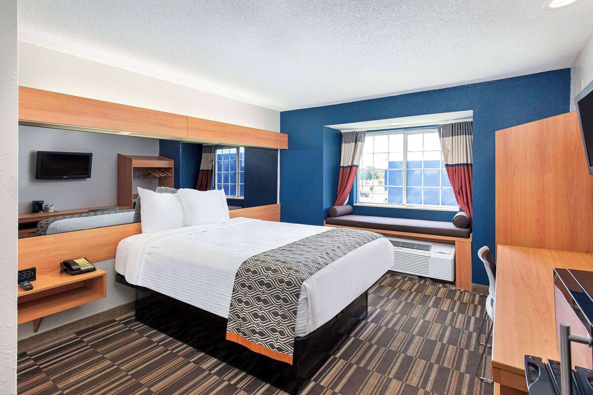 A hotel room with a bed, a desk, a chair, a TV, a window with curtains, and a view of the outside.