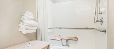 Ducha con barra de apoyo, banco y una pila de toallas en la pared.