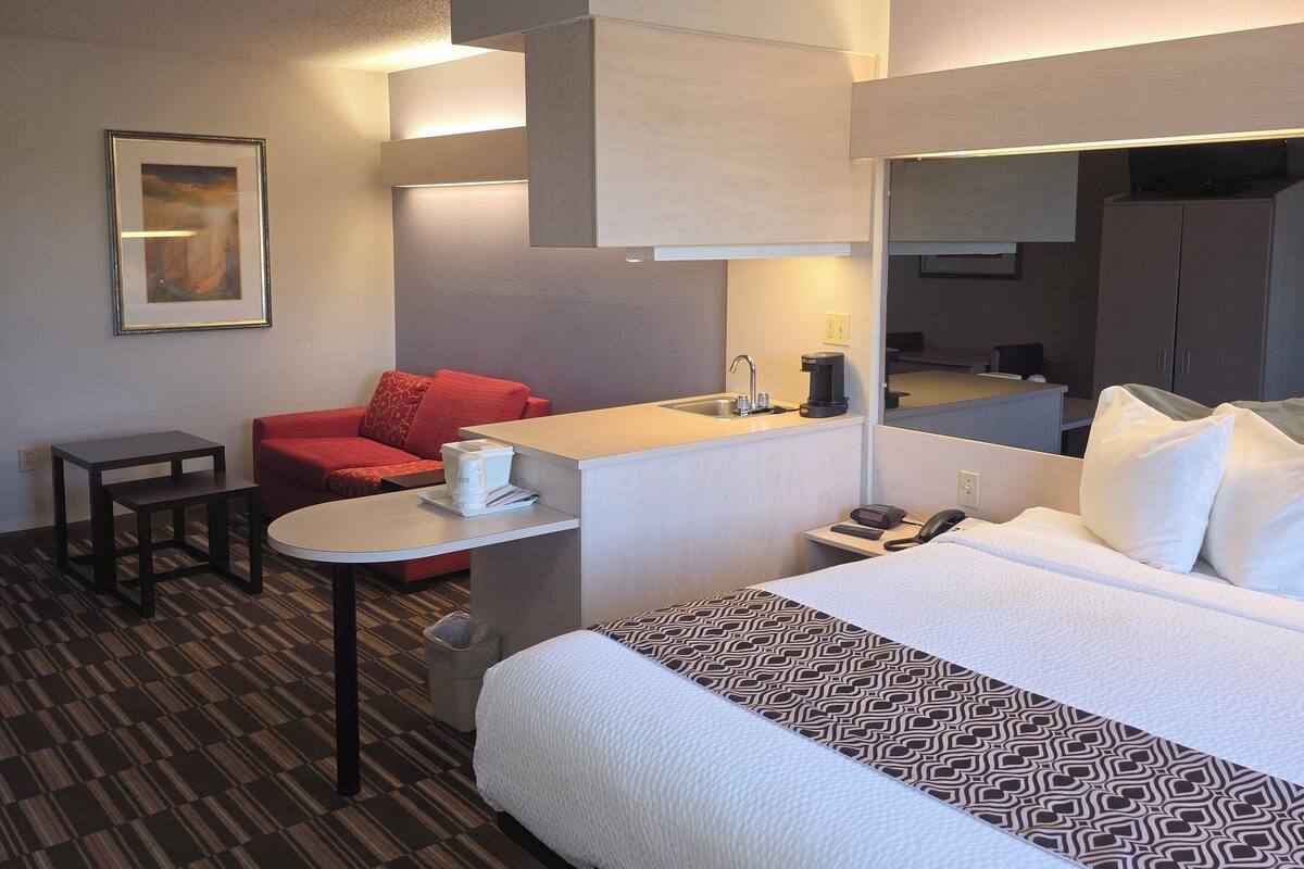 A hotel room with a bed, a desk, a chair, a sofa, a coffee machine, a phone, and a painting on the wall.