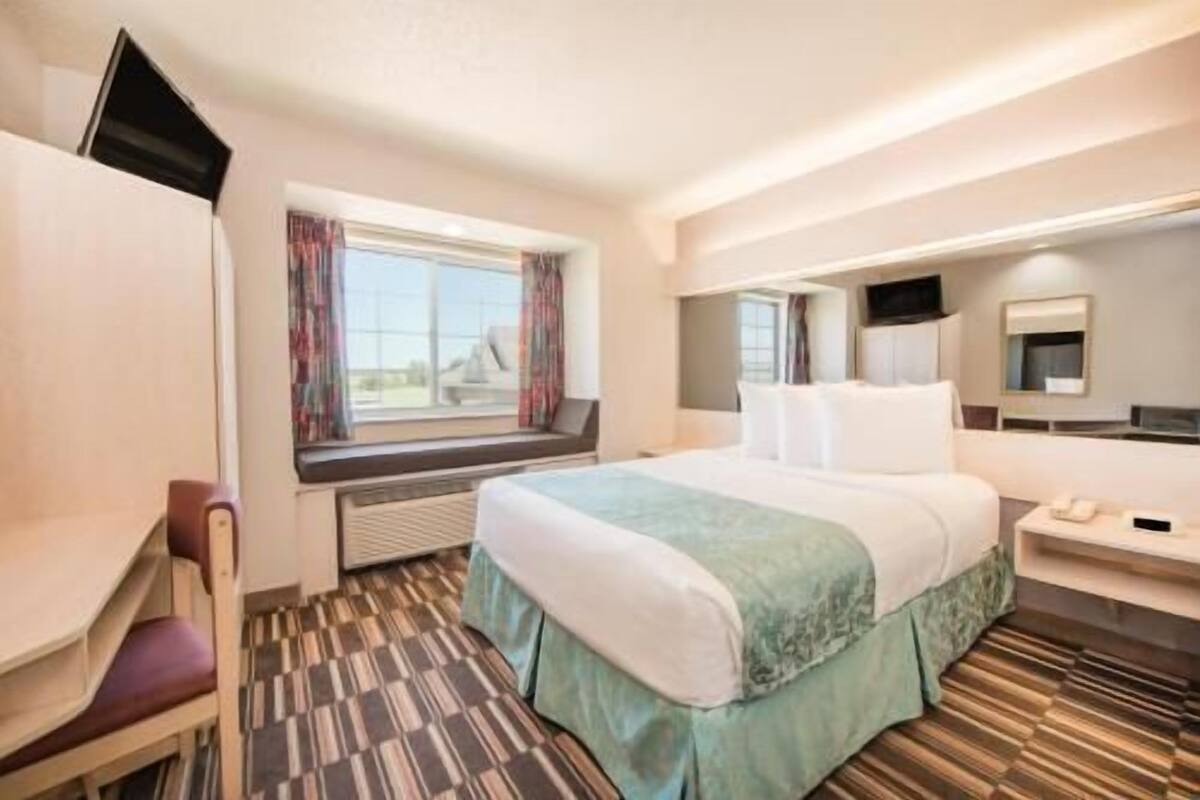 A hotel room with a bed, a desk, a chair, a window with curtains, a television, and a mirror.
