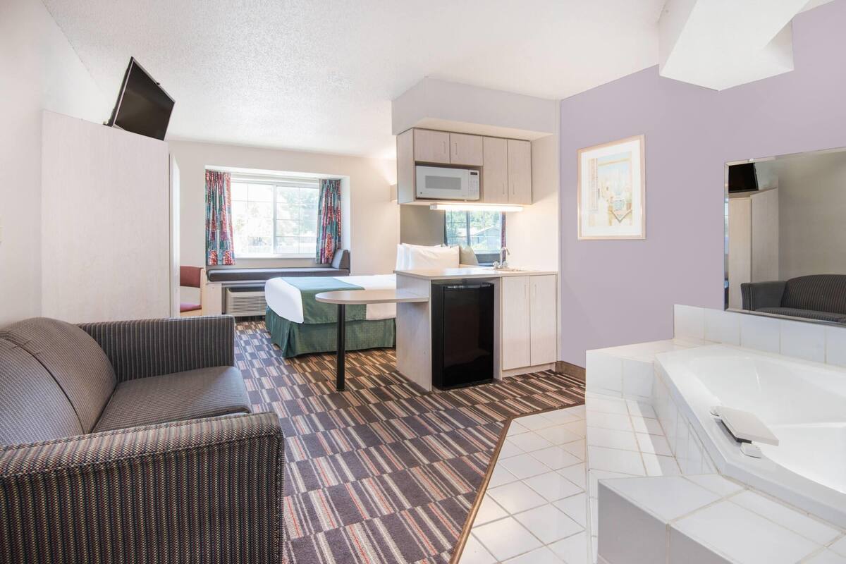 A compact room with a sofa, a small kitchenette, a window with curtains, a white bathtub, and a flat-screen TV mounted on the wall.