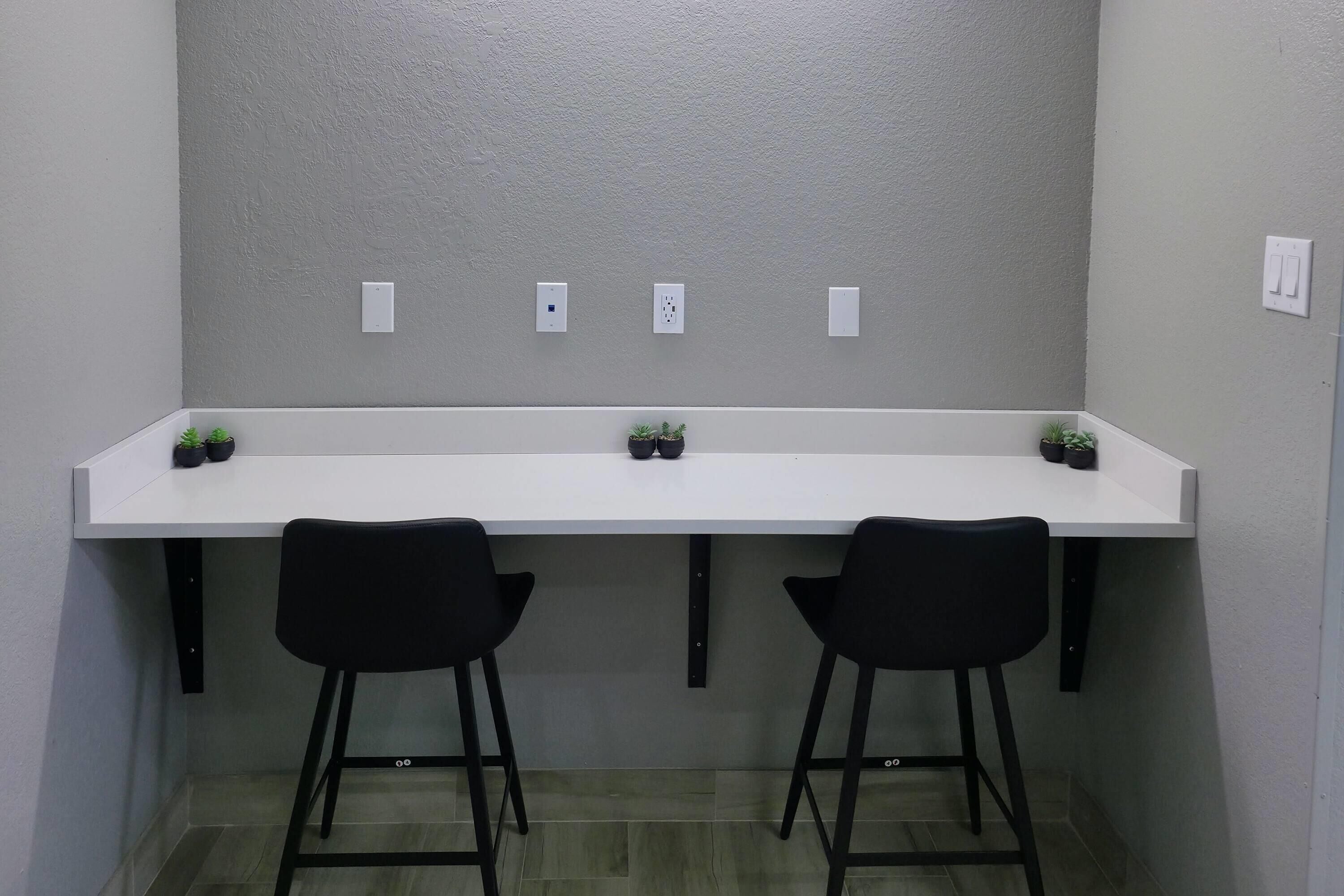Un escritorio blanco con dos sillas negras, dos plantas pequeñas en macetas y tres enchufes en la pared.