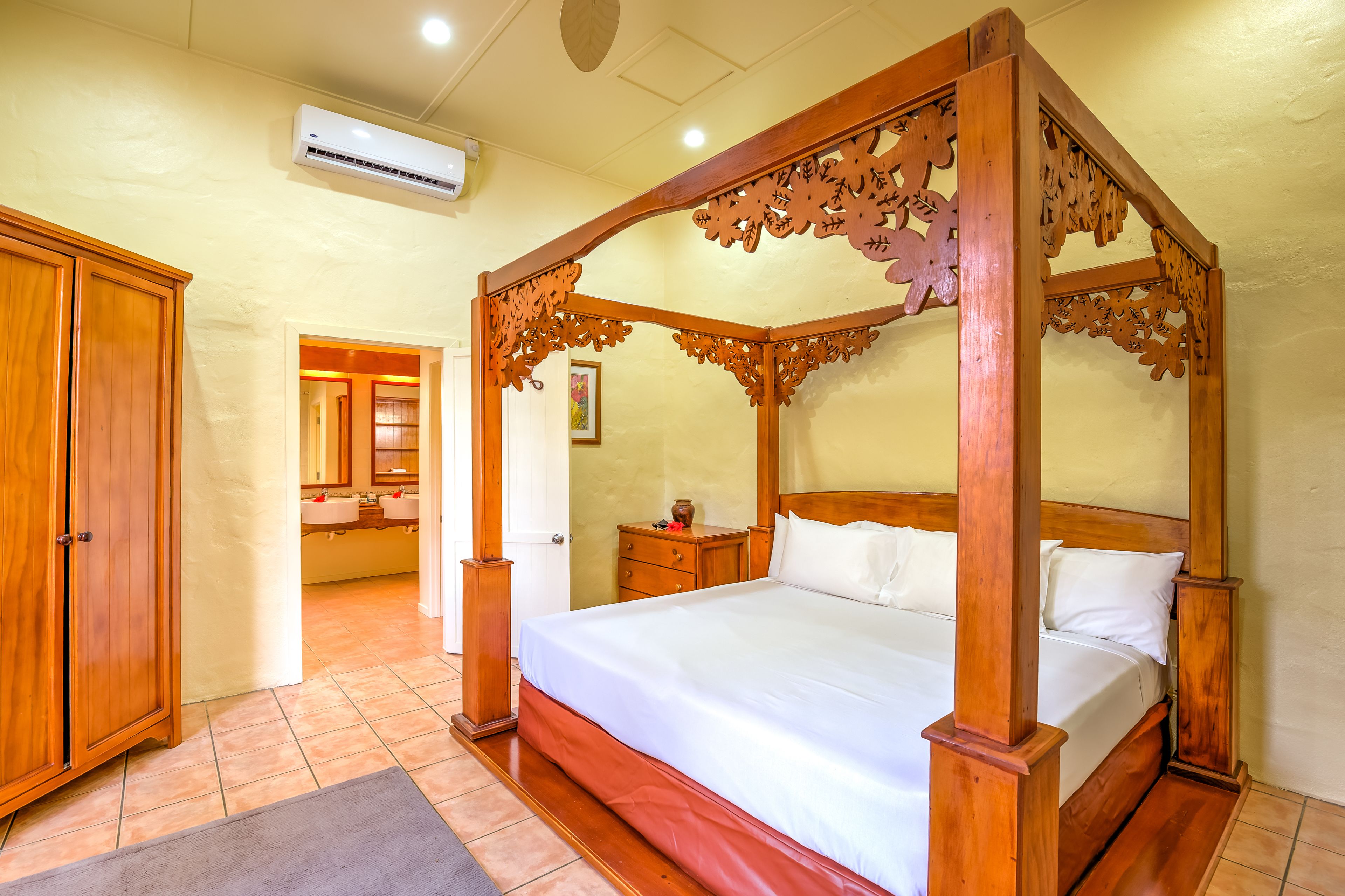A four-poster bed with a wooden frame and white bedding, a wooden wardrobe, and a bathroom with a sink and mirror.