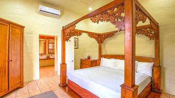 A four-poster bed with a wooden frame and white bedding, a wooden wardrobe, and a bathroom with a sink and mirror.