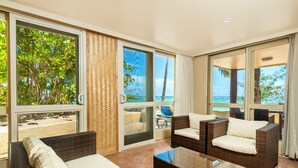 Une pièce spacieuse dotée d’une grande porte-fenêtre coulissante qui donne sur un balcon offrant une vue sur la plage et l’océan. On y trouve deux canapés en osier et une table basse en verre.