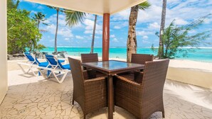 Un espace extérieur aménagé, abrité et doté d’une table en verre et de chaises en osier, offrant une vue sur une plage bordée de palmiers et baignée d’une eau turquoise.