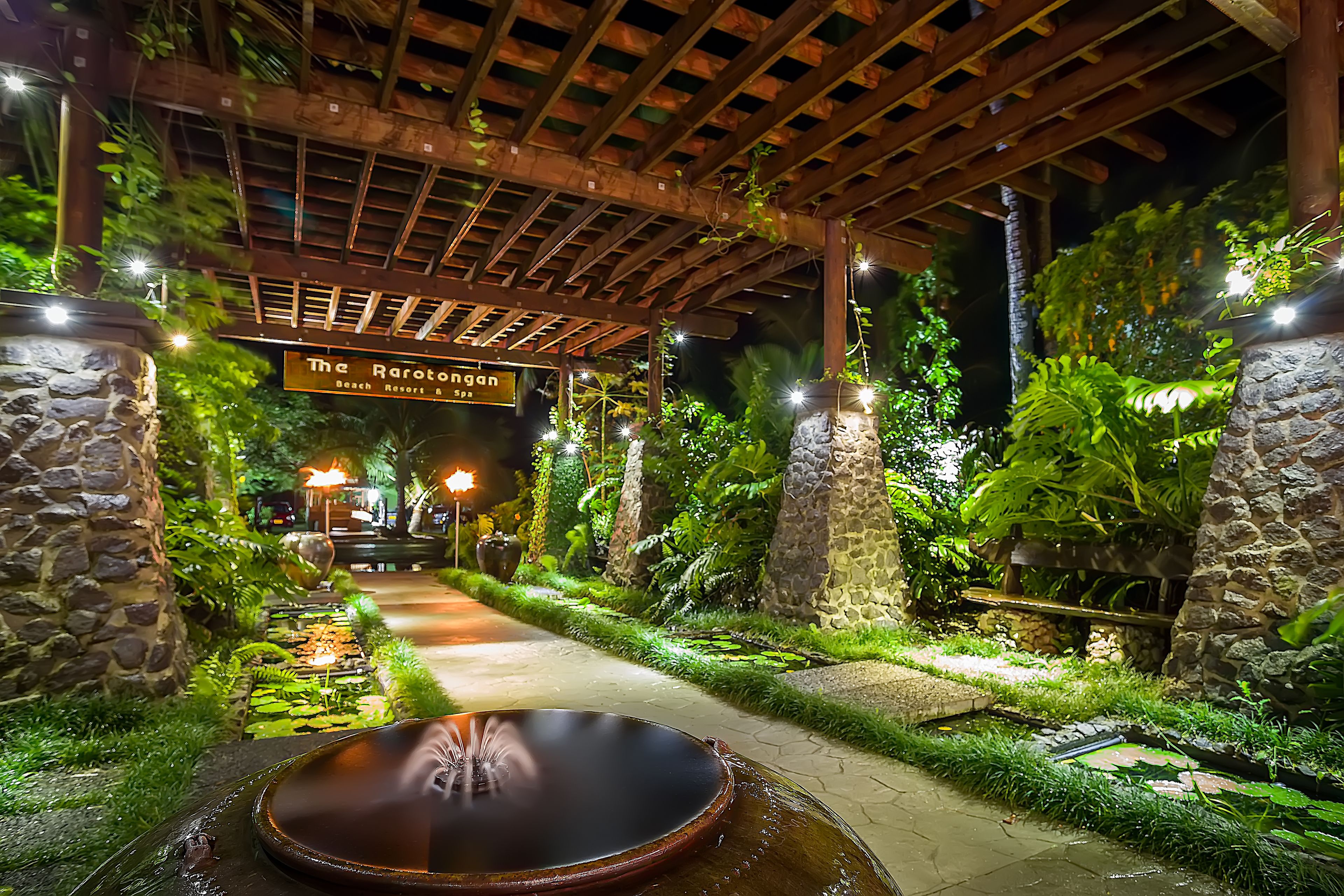 A well-lit pathway leading to a spa area with a hot tub, surrounded by lush greenery and stone walls.