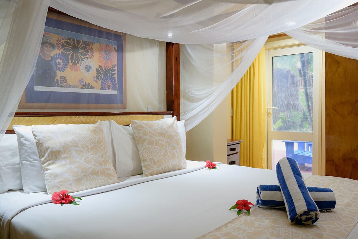 A neatly made bed with white linens and decorative pillows, a canopy overhead, and a tropical-themed painting on the wall.