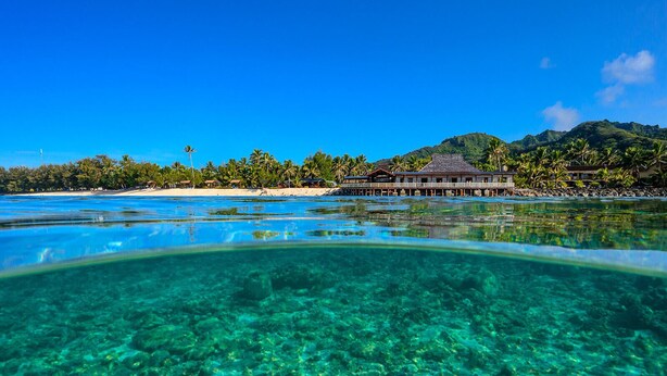 On peut clairement voir un complexe hôtelier avec des bungalows sur pilotis, une plage et une chaîne de montagnes en arrière-plan.