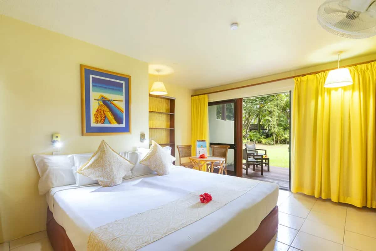 A hotel room with a bed, a painting, a small table with chairs, and a view of the outdoors through a sliding door.