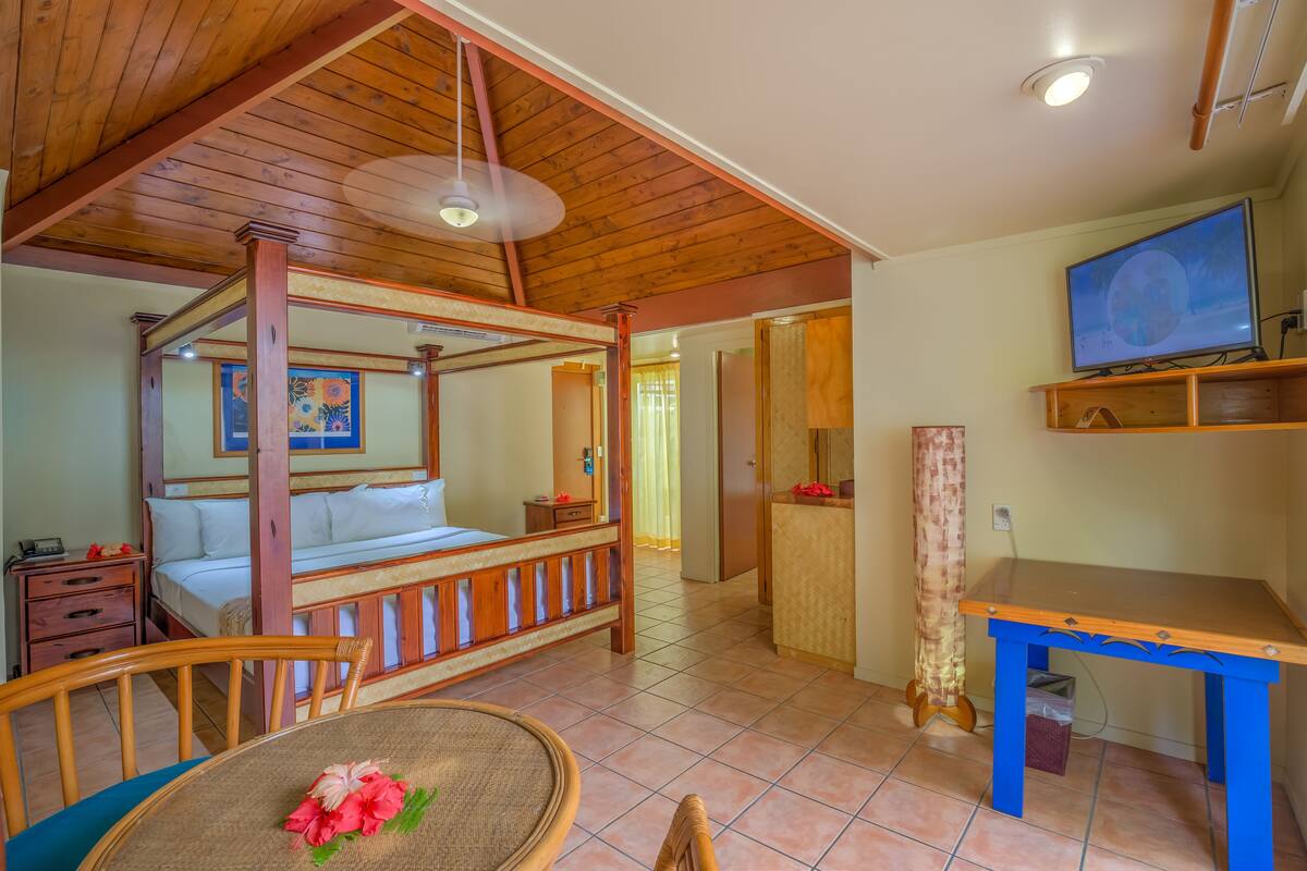 A room with a four-poster bed, a wooden ceiling, a television on a shelf, a table with a chair, and a small desk.