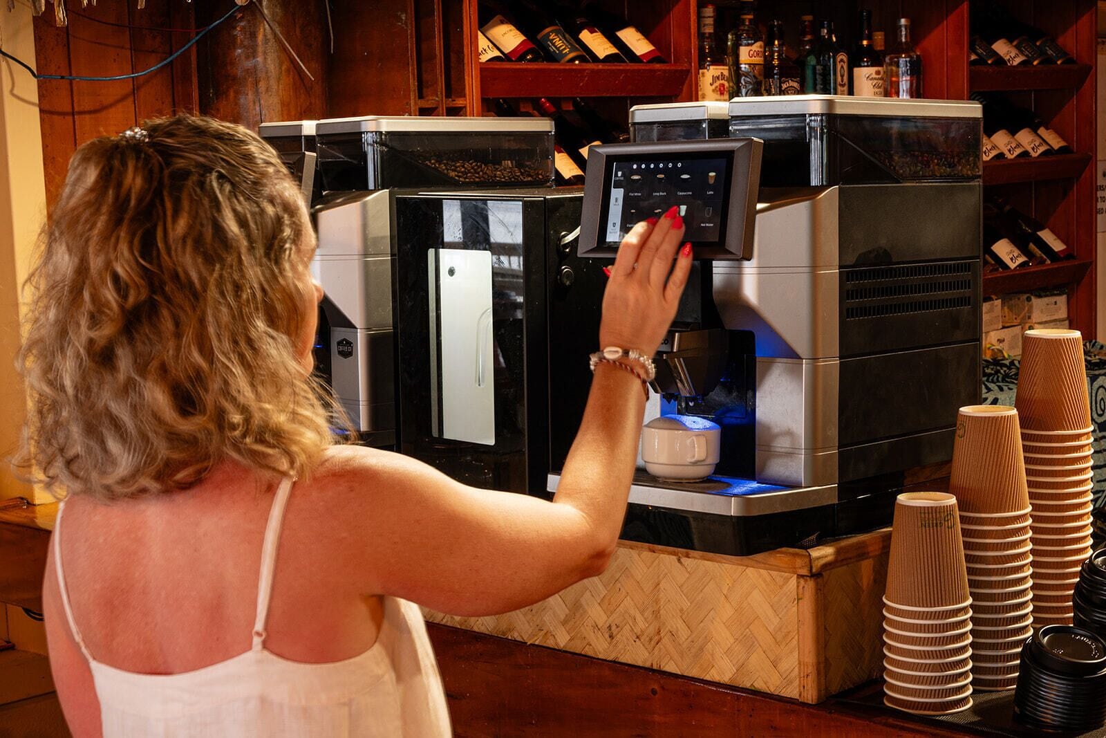 Una persona operando una máquina de café con pantalla digital, rodeada de pilas de vasos de papel y una torre de vinos al fondo.