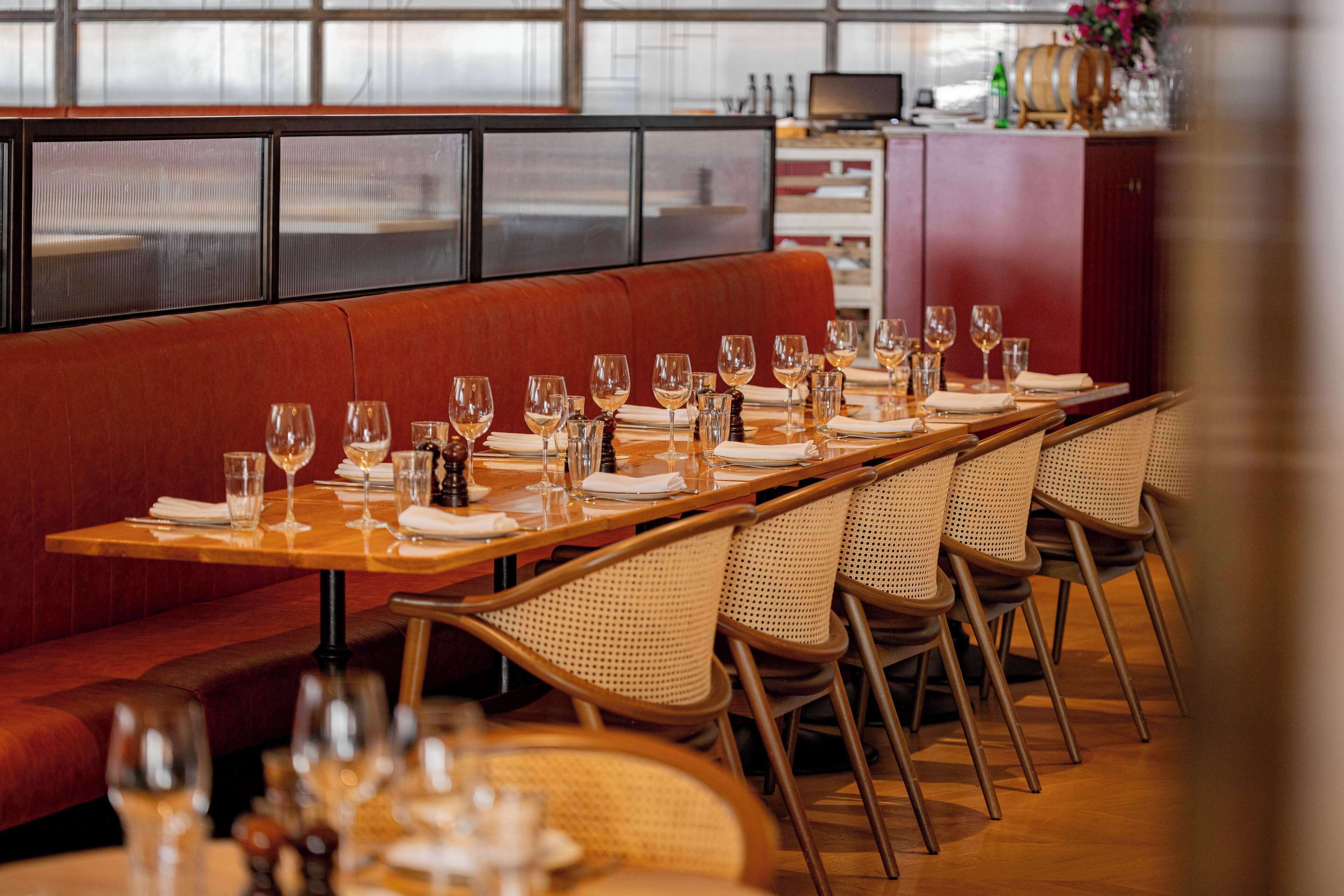 A restaurant with a long wooden table set for a meal, wicker chairs, and a red cushioned bench.