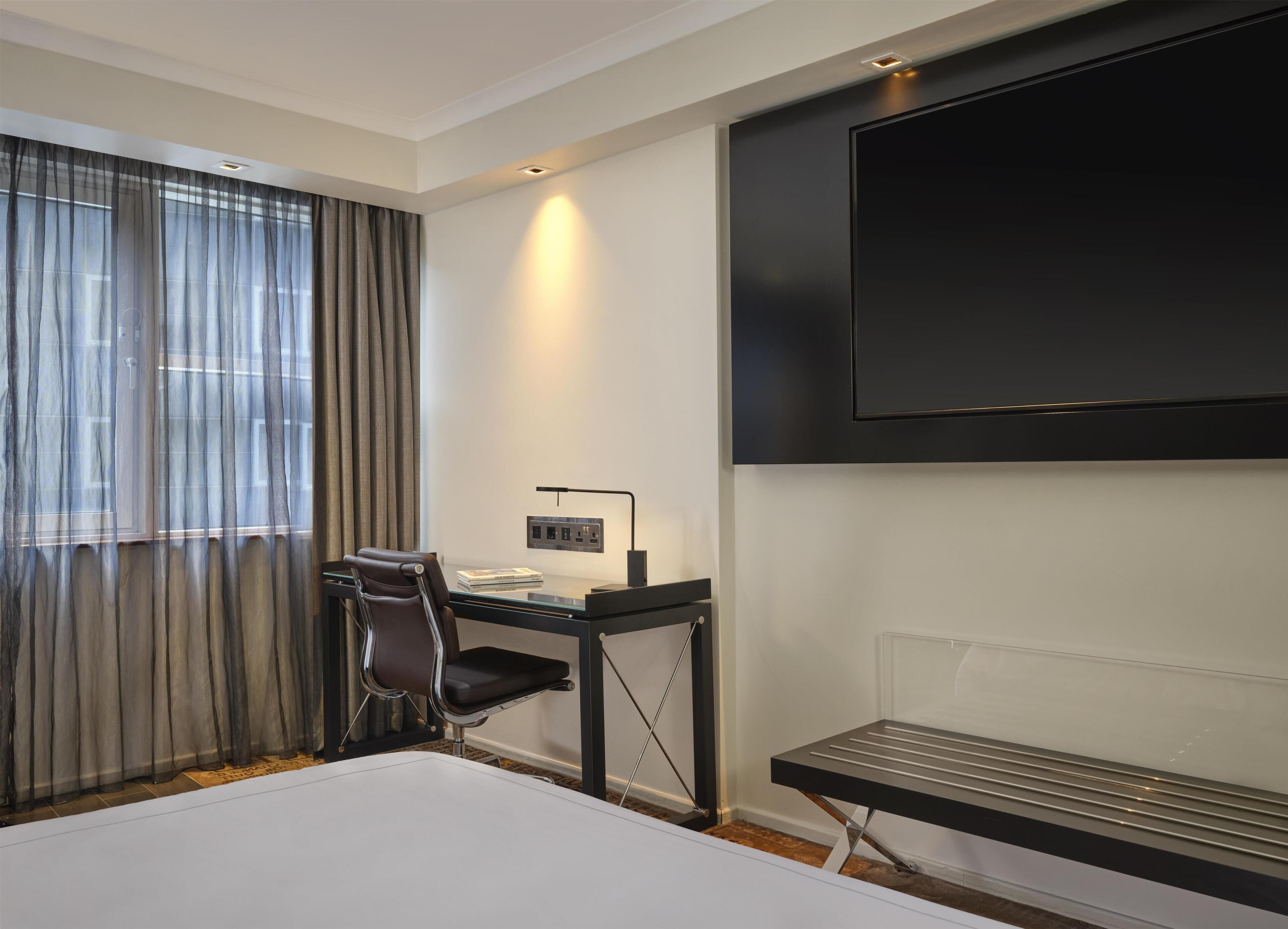 Habitació d'hotel amb llit, escriptori, cadira, televisió i finestra amb cortines.